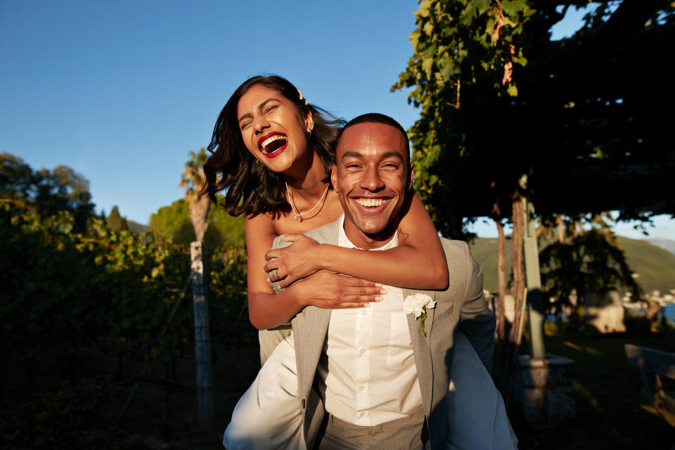 Man in a suit giving piggyback ride to woman in dress, both laughing, outdoors