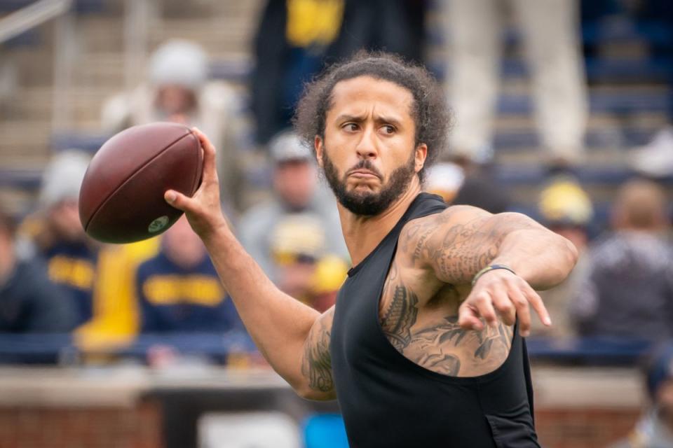 A Black man wearing a black tanktop holds a football in his left hand, looking like he is about to throw it. People sit in the stands behind him. 