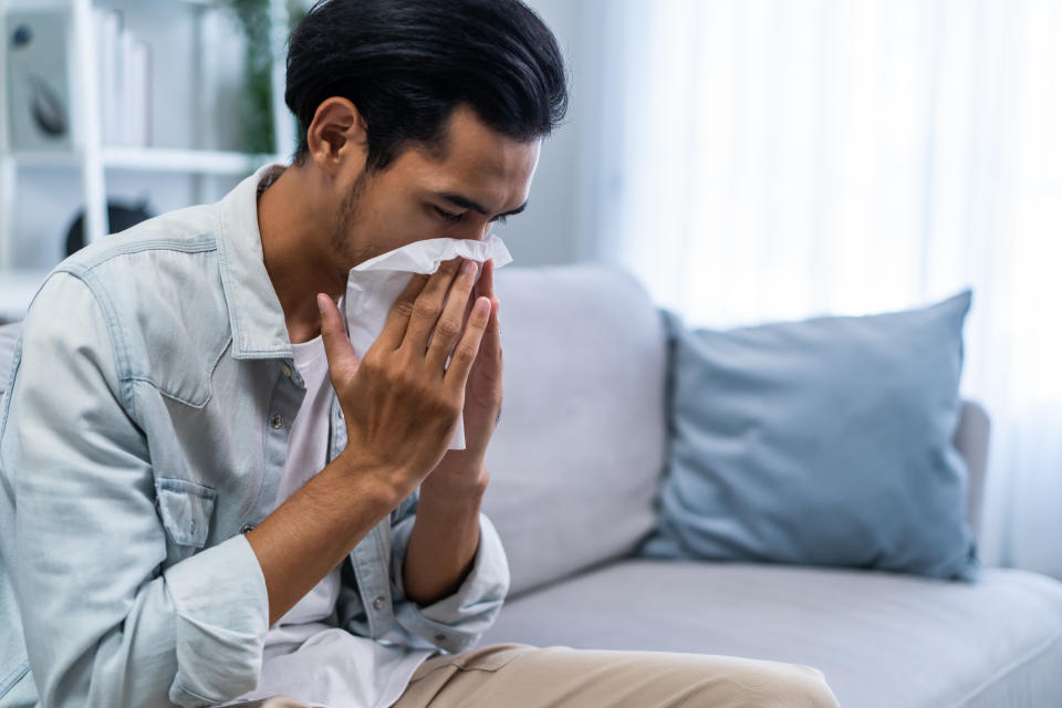 Asian handsome male having running nose and sneezing in living room. Attractive young man sitting on sofa feeling bad and suffer from allergy then put tissue cover her nose while sneez in house.