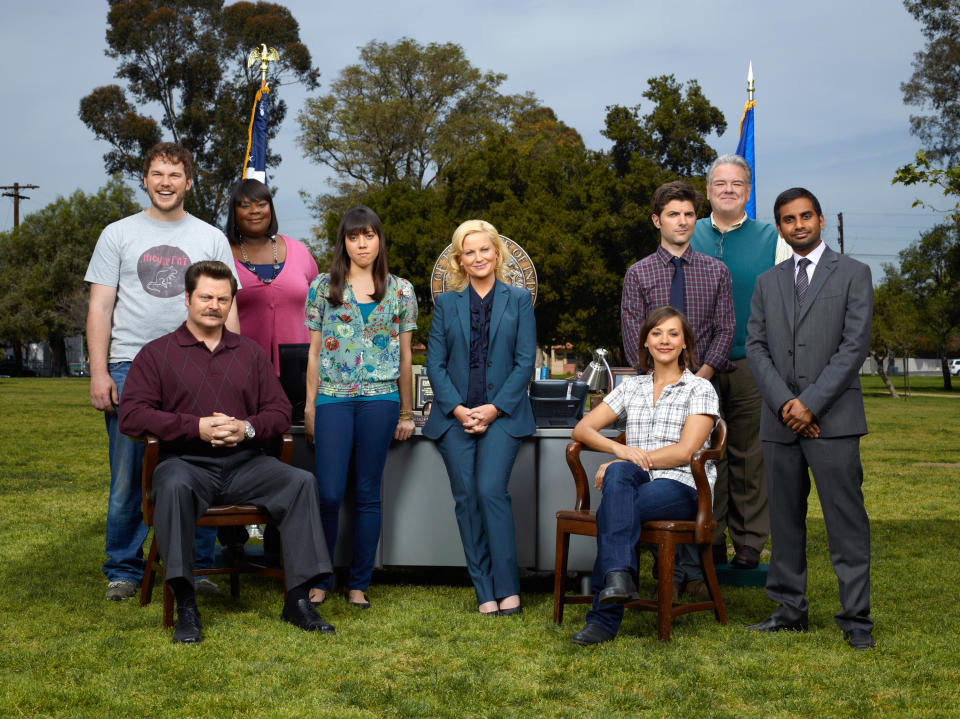 The cast of Parks and Rec standing or sitting in chairs on grass