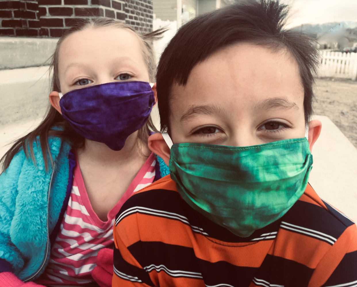 Two children, a Caucasian boy and a Caucasian girl, sitting outside wearing fabric home-made face masks to cover their nose and mouth. You can see that they are smiling beneath their masks.