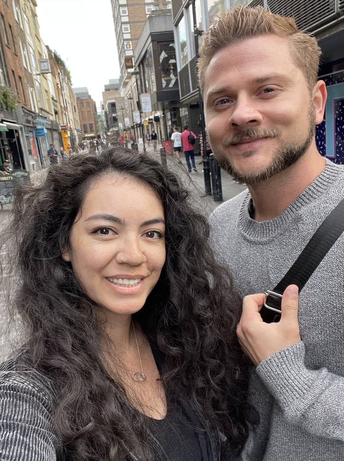 Two people, a woman with long curly hair and a man with short hair and a beard, smile while standing on a city street