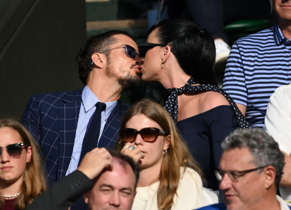 Orlando Bloom and Katy Perry share a kiss, both dressed in smart attire at an outdoor event