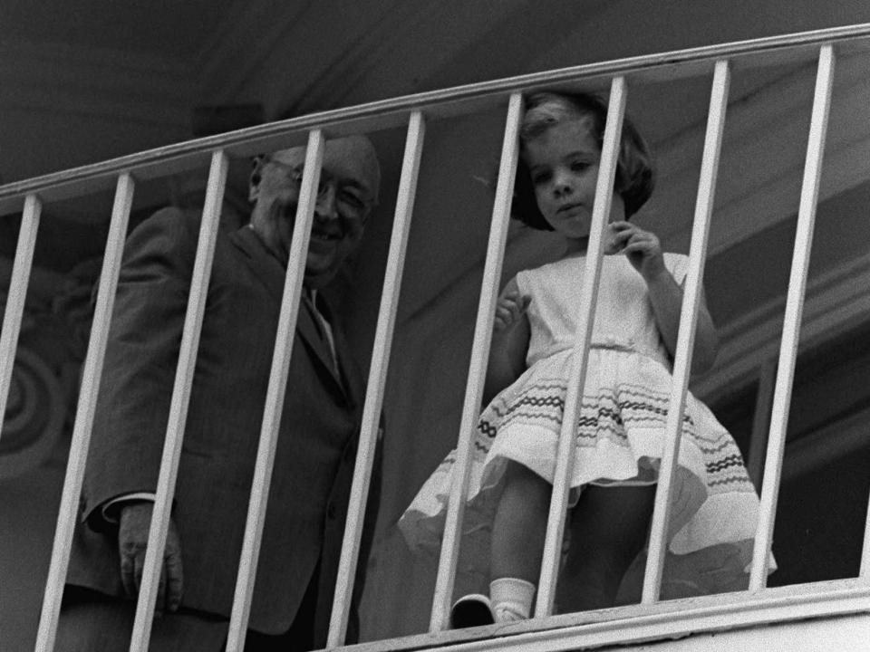 Caroline Kennedy peers through the bars of a balcony as President John F. Kennedy and Jackie Kennedy host a garden party at the White House.