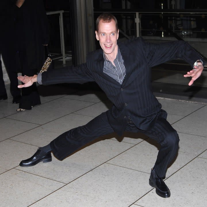 Doug Jones does a one leg squat in a black striped suit