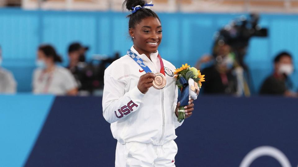 simone biles stands and looks to the right, she holds an olympic medal in one hand that is hanging around her neck and a flower bouquet in the other hand, she wears an all white athletic suit and a blue and white hair ribbon