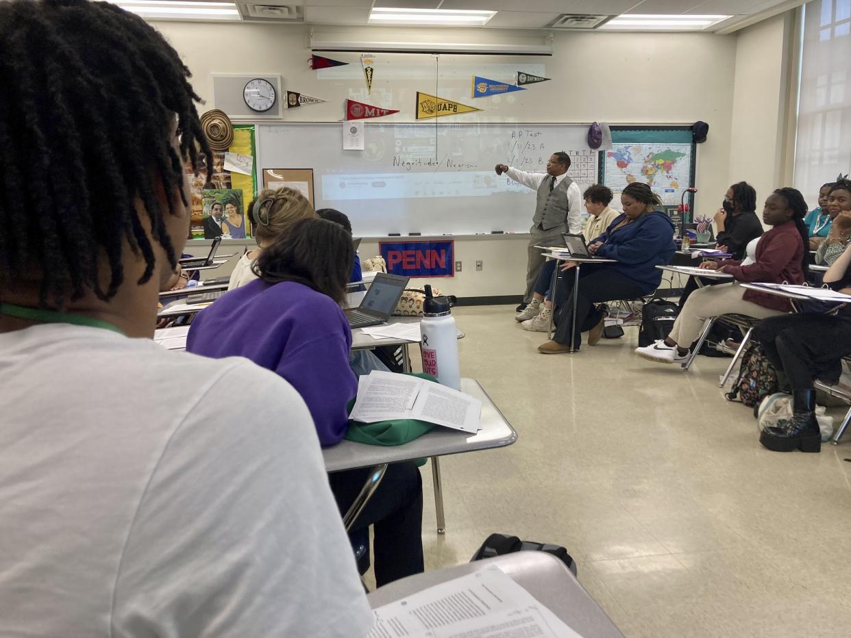 A teacher stands at the front of the classroom and points to a white board as students look on.