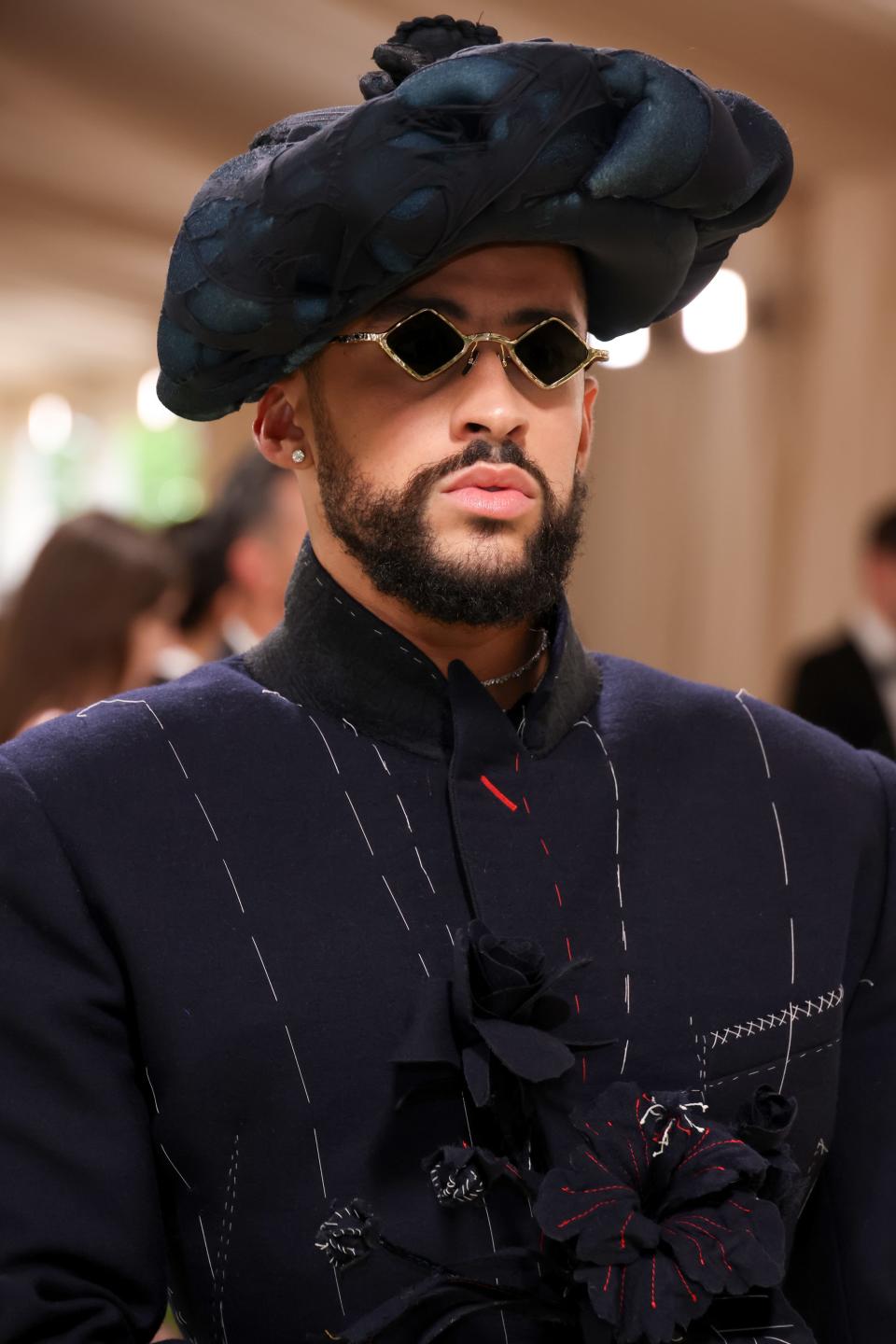 Bad Bunny wearing a sculptural hat, sunglasses, and a dark jacket with line patterns and floral adornment