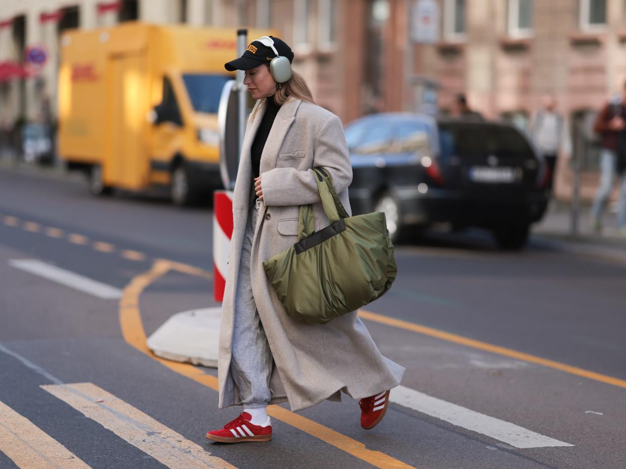 Sonia Lyson is seen wearing a black Sporty & Rich cap, silver Apple AirPods Max headphones, black turtleneck sweater from Zara, grey Weekday jogger pants, beige long wool coat from Edited, green VeeCollective shopper bag, Balenciaga white logo socks and red Adidas Gazelle sneakers, on October 07, 2022