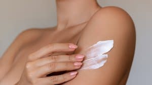 Cropped shot of a young tanned woman moisturizing her shoulder with her hand using a cream isolated on a gray background. Skin care. A white thick smear of cream on the female body