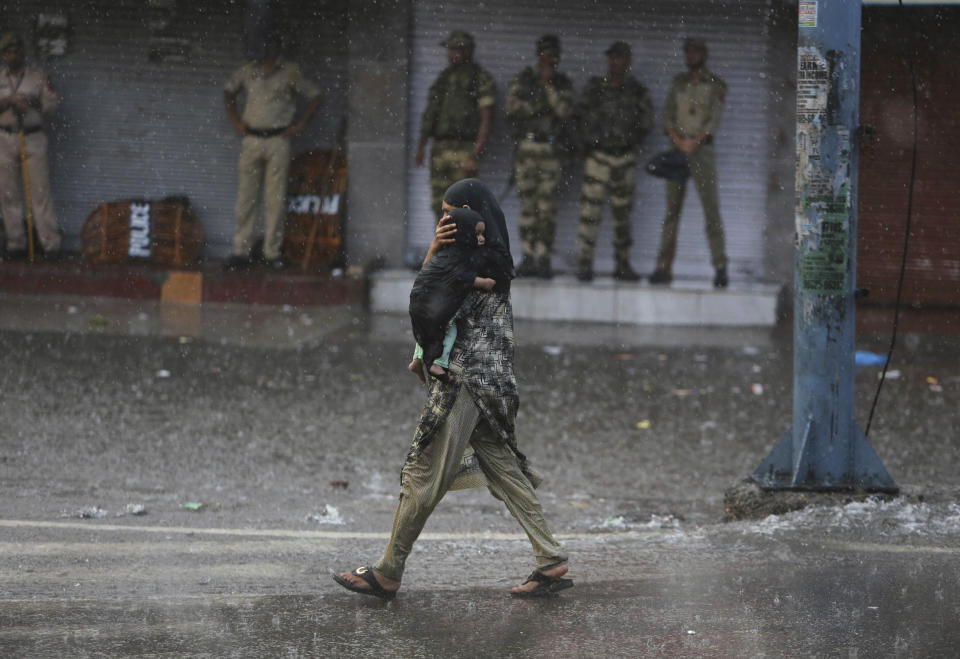 A woman carries her child as she walks past Indian security personnel as it rains during curfew like restrictions in Jammu, India, Monday, Aug. 5, 2019. An indefinite security lockdown was in place in the Indian-controlled portion of divided Kashmir on Monday, stranding millions in their homes as authorities also suspended some internet services and deployed thousands of fresh troops around the increasingly tense region. (AP Photo/Channi Anand)