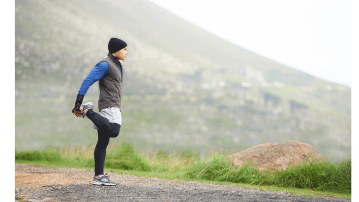 Man standing on a rural road near foothills standing on one leg with the other heel bent back toward his glutes to stretch his quads