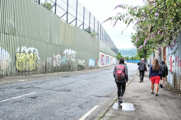 People walk on a pavement past a tall corrugated iron barrier.