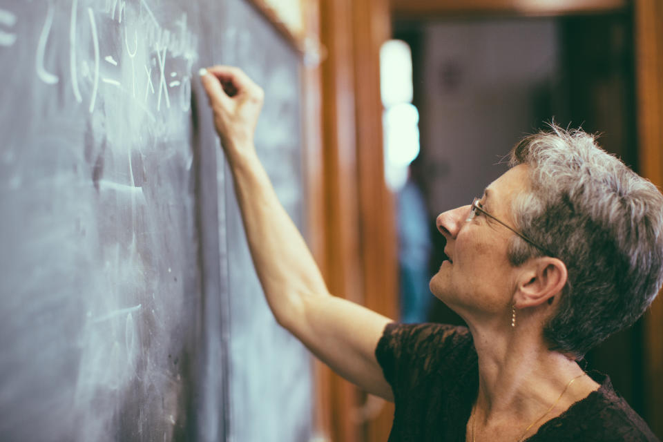 A person with short hair and glasses writes mathematical equations on a chalkboard in a classroom