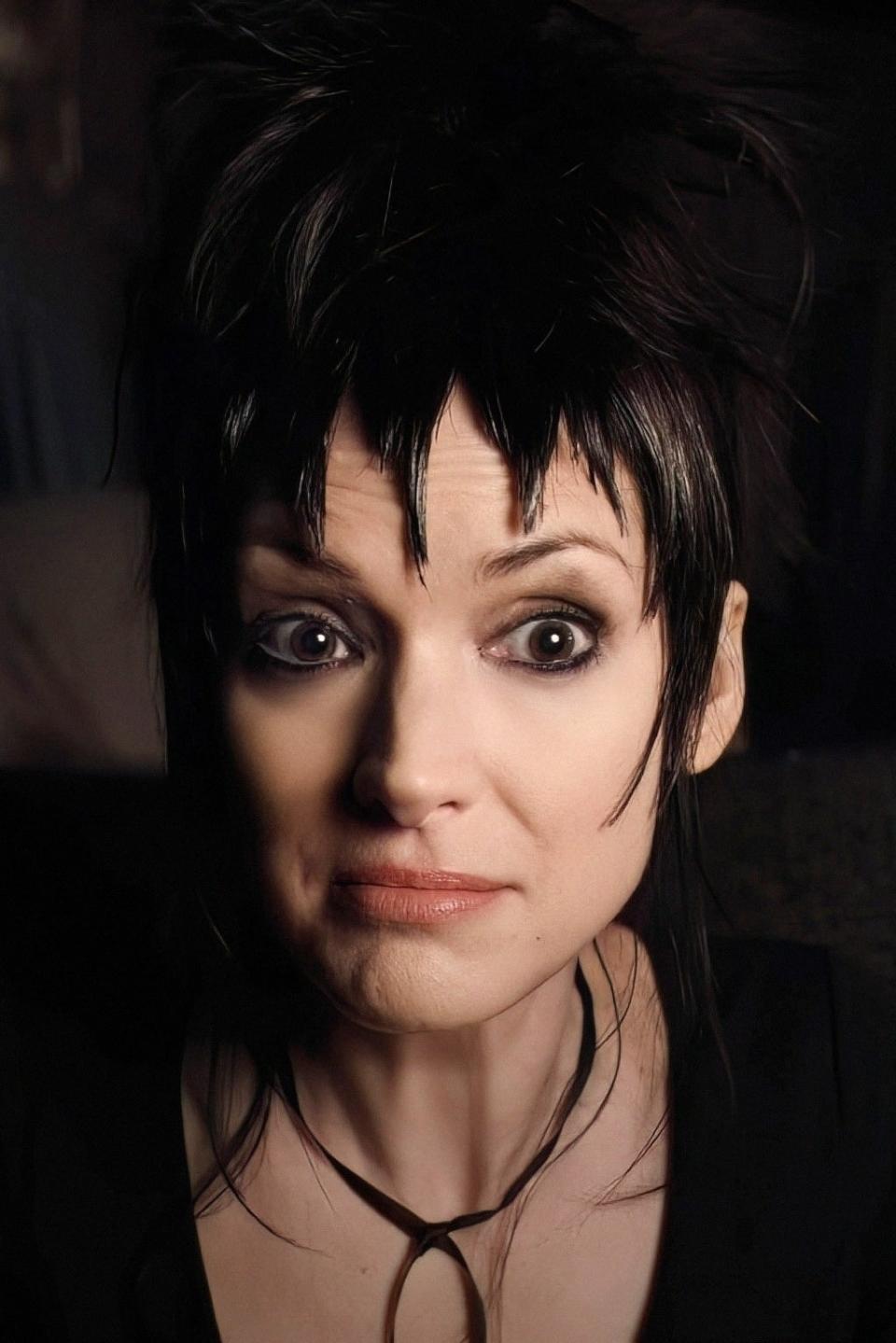 Winona Ryder with short, spiky black hair, wearing a black top and sitting in a dimly lit room