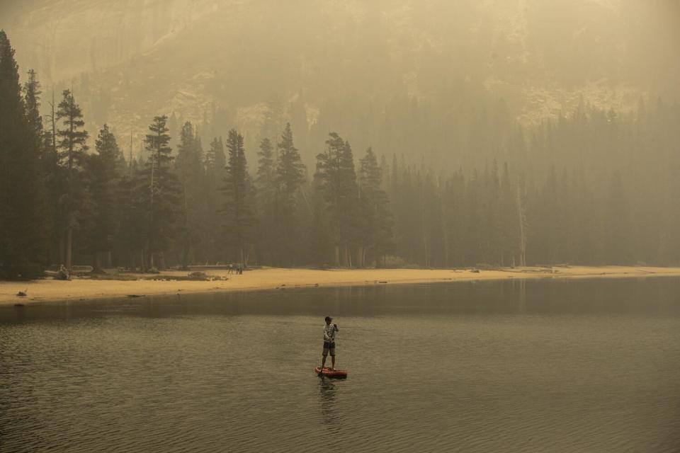 A lone figure paddle boards on a smoke-shrouded lake.