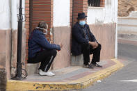 Two men wearing face masks to protect against the spread of coronavirus sit and talk on a street corner in the rural village of La Puerta de Segura, Jaen, Spain, Tuesday, Oct. 27, 2020. Despite overnight curfews, mobility curbs affecting at least four regions and other restrictions, health authorities in Spain are warning that sharp outbreaks of Covid-19 are already straining hospitals. (AP Photo/Paul White)