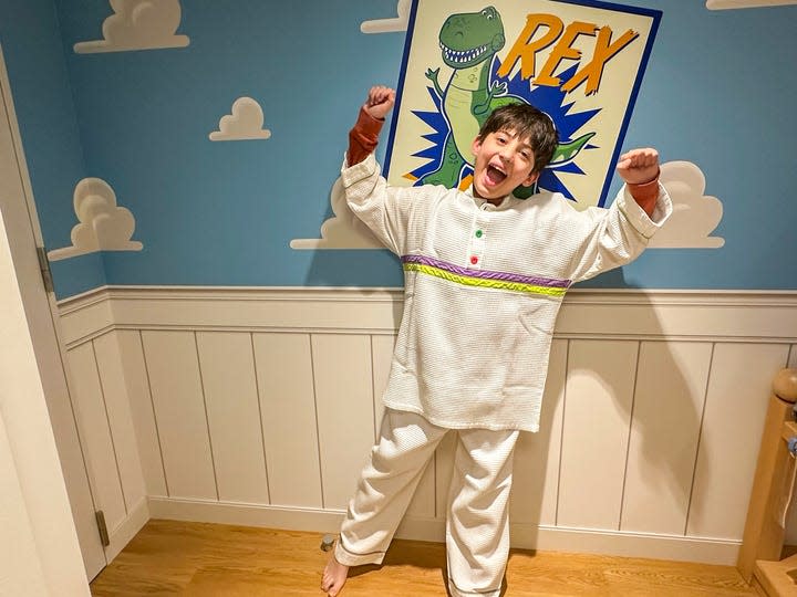 A child wears a set of pajamas included with a stay at the Toy Story Hotel. He poses in front of a dinosaur poster with his arms raise and a smile on his face.