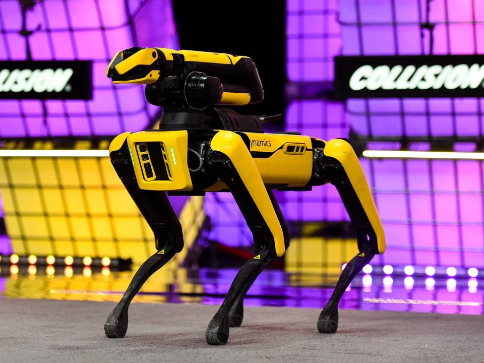 A yellow quadrapedal robot with a mechanical arm attached to its back stands on a stage in front of a purple backdriop that says "collision."