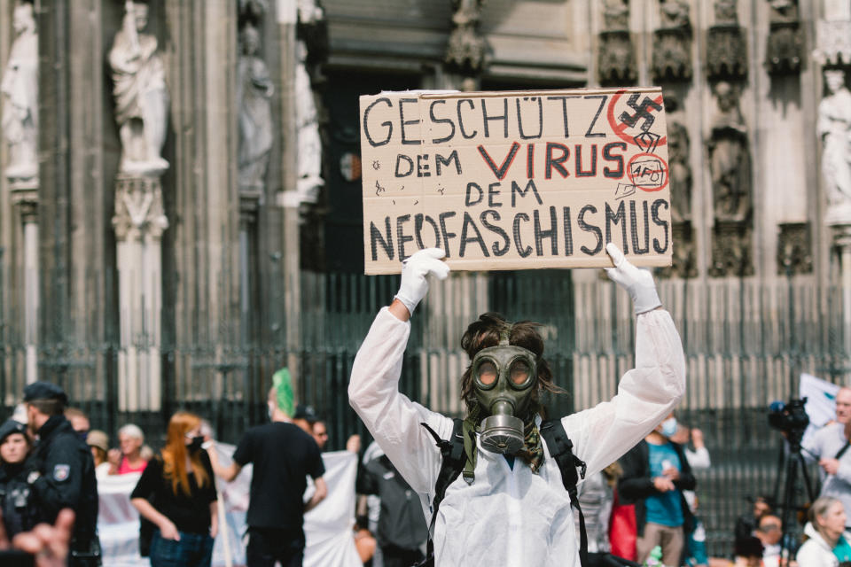 Un hombre con una máscara antigás y un traje protector sostiene una pancarta con el mensaje “protégete del virus nazi”, durante la manifestación contra las políticas para paliar las consecuencias del coronavirus en Colonia, Alemania, el 16 de mayo de 2020 (Ying Tang / NurPhoto).