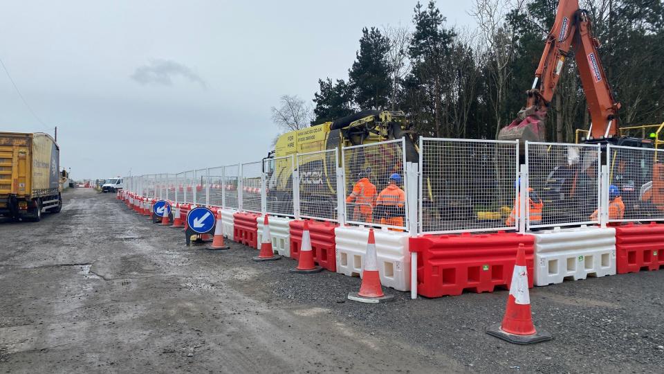 Workers with diggers behind a metal fence with a dirt road next to it 