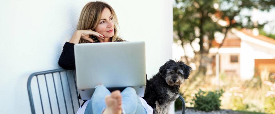 Woman and dog sit with laptop on bench outside home