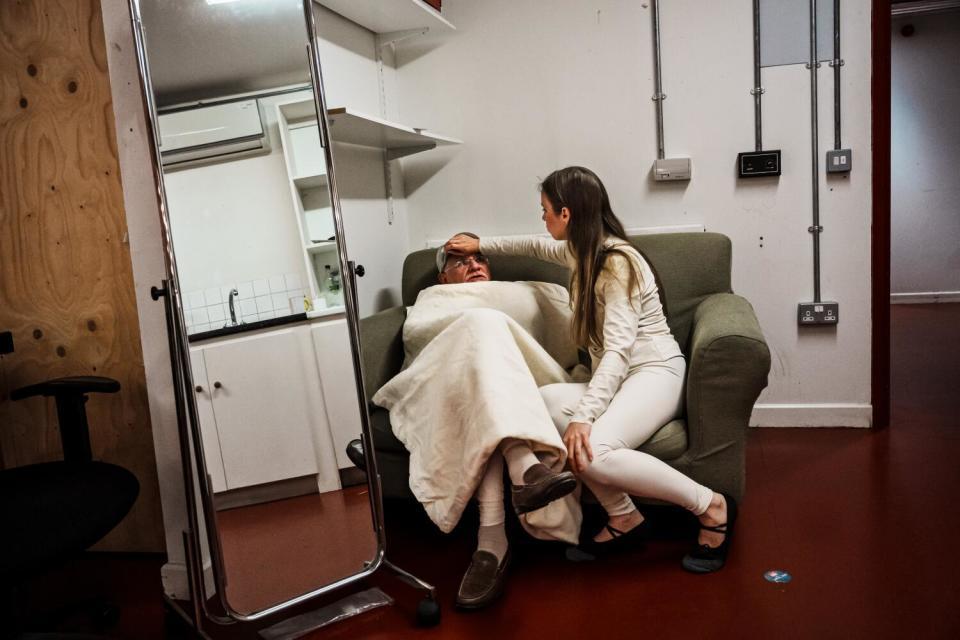 A woman puts her hand on the forehead of a man covered with a blanket and seated next to her on a couch