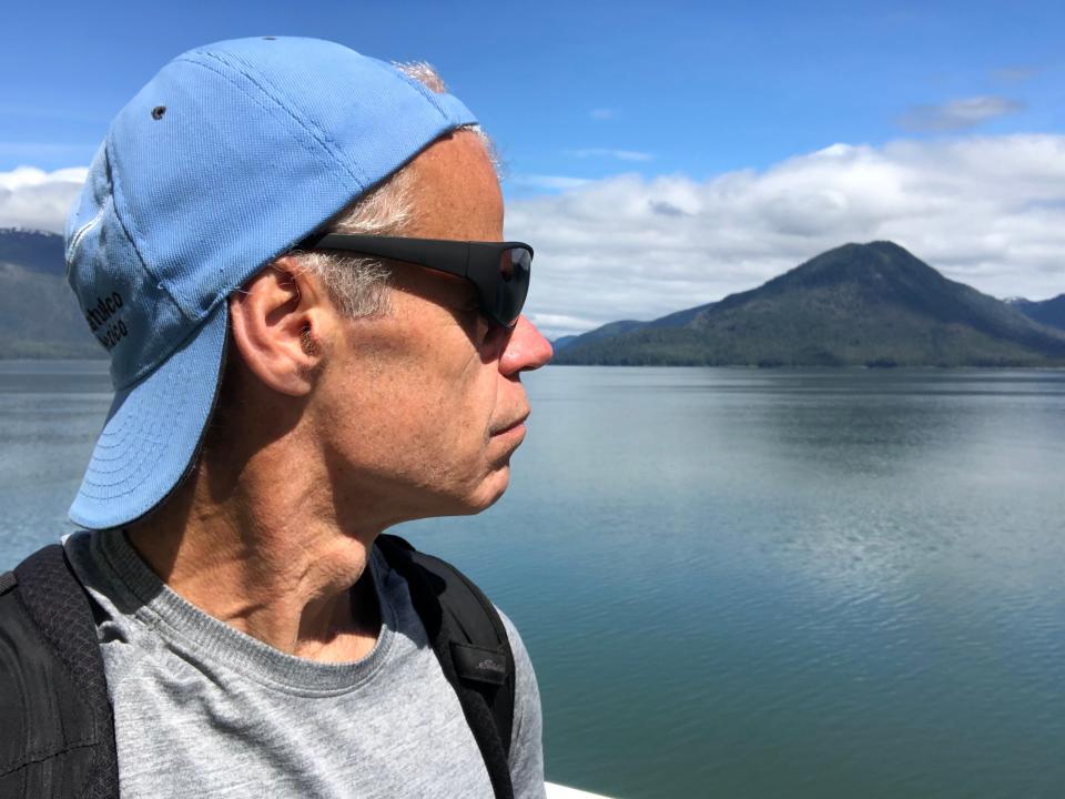 The profile of a man with a backwards blue baseball hat wearing sunglasses with an island and water in the background.