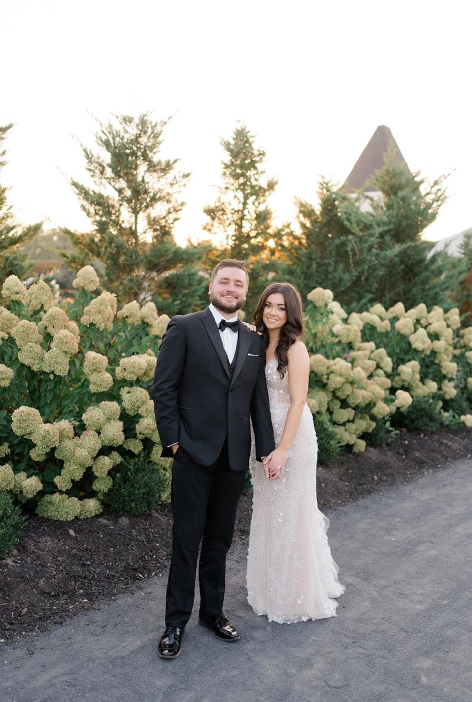 A bride and groom hold hands in front of white hydrangea bushes.