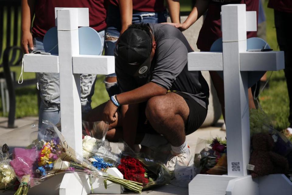 A man crouches next to a row of small white wood crosses, his head down as others behind him lay their hands on his shoulders. Bouquets of flowers lay on the ground.