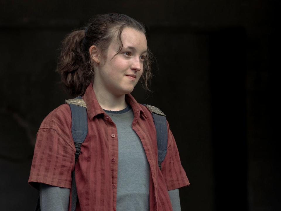 bella ramsey as elli on the last of us, wearing a slate grey shirt and red overshirt and looking fondly to the side