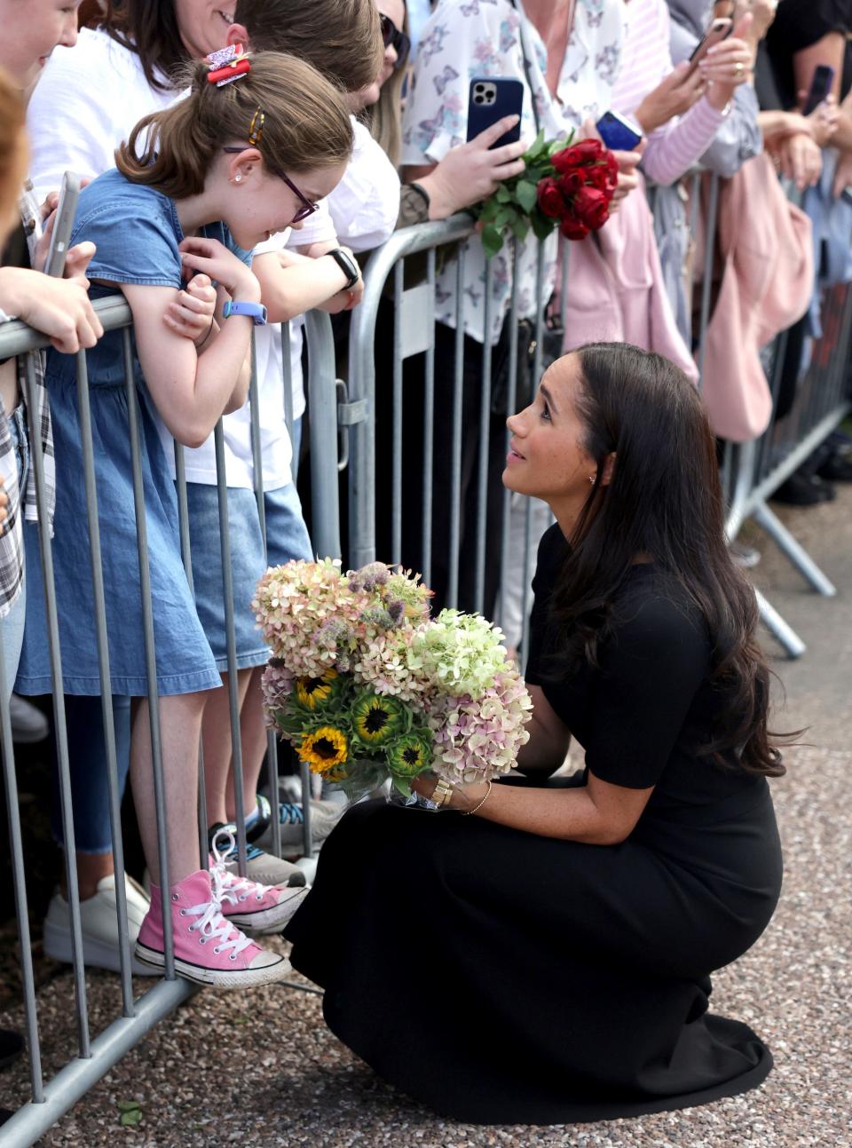 Meghan Markle kneels holding flowers to speak to a little girl who leans over a fence.