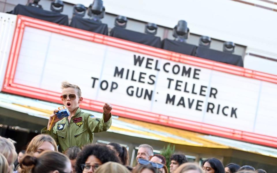 a boy in a flight suit in front of a marquee for top gun maverick maverick could be a popular baby boy name in 2023 fitting in with the neo cowboy name trend