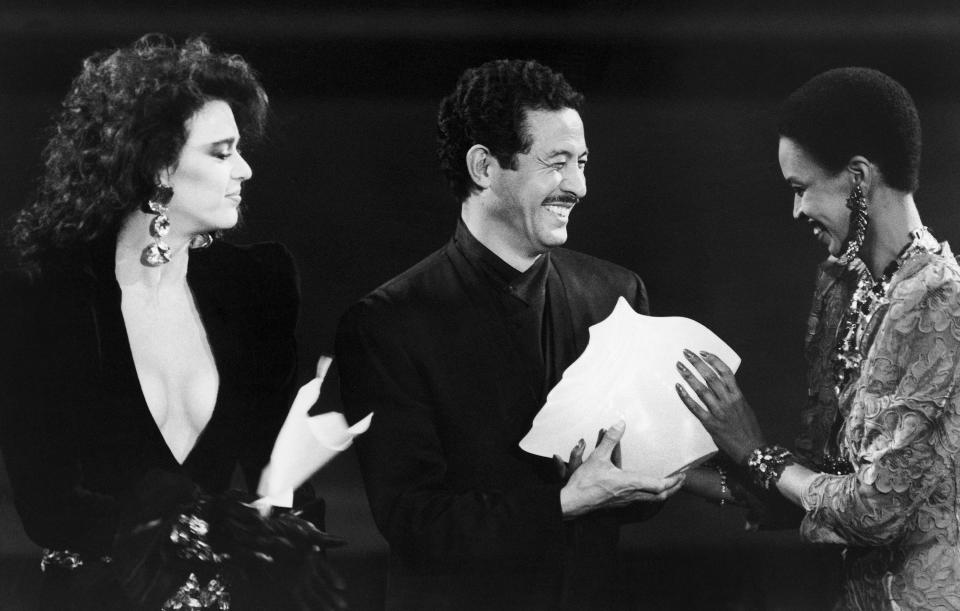 FILE - Japanese fashion designer Issey Miyake receives the award for Showing Best Paris Collection by a Foreigner from an unidentified model, while he is applauded by Dutch actress Maruschka Detmers, left, during the "Oscars" for fashion award ceremony at the Paris Opera, France, on Oct. 24, 1985. Miyake, who built one of Japan’s biggest fashion brands and was known for his boldly sculpted pleated pieces as well as former Apple CEO Steve Jobs’ black turtlenecks, has died. He was 84. He died Aug. 5, 2022, of liver cancer, Miyake Design Office said Tuesday, Aug. 9. (AP Photo/Michel Lipchitz, File)