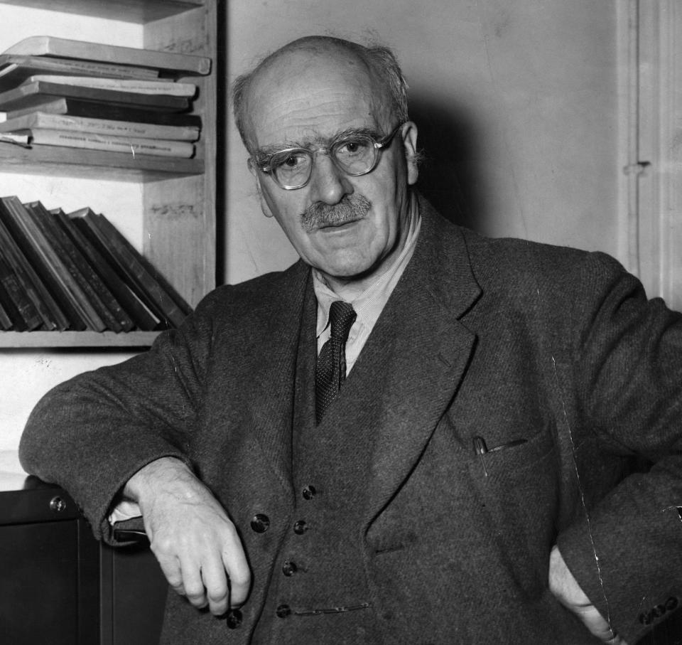 JBS Halane wearing glasses and with a mustache and wearing a suit stands next to a bookshelf in an office