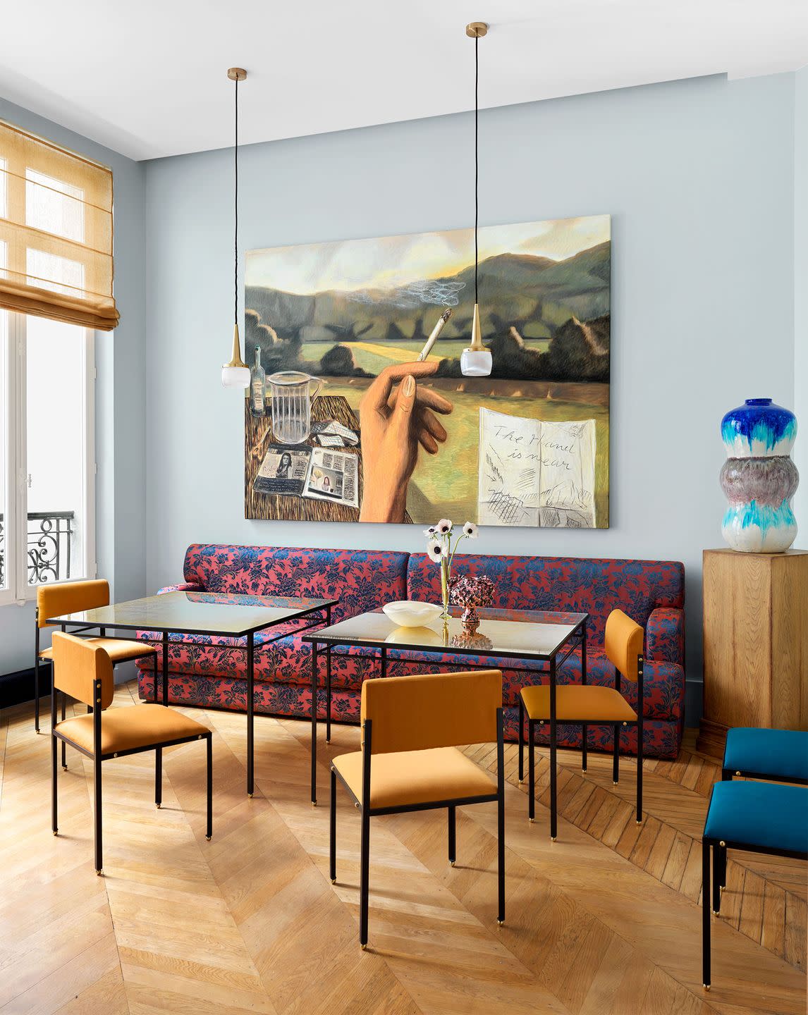 a dining room has light blue walls, a rose and deep blue patterned banquette, two square glass topped tables, four orange and two blue dining chairs, twin frosted glass pendants, and a surrealist painting,