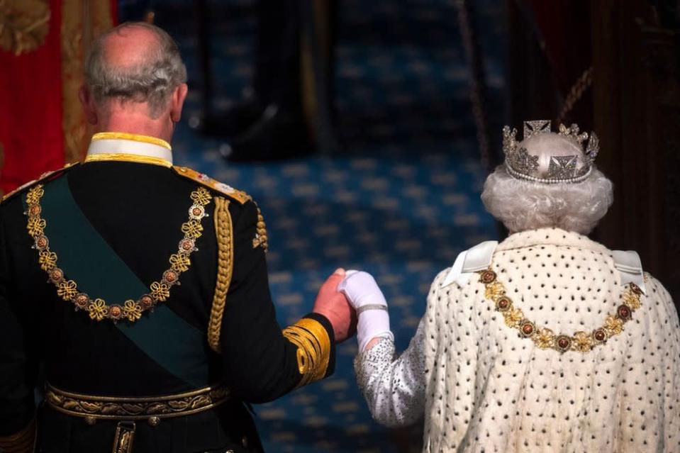 Fotografia colorida mostra a rainha de costas com um manto cerimonial e uma coroa, segurando a mão de Charles, também de costas de uniforme militar