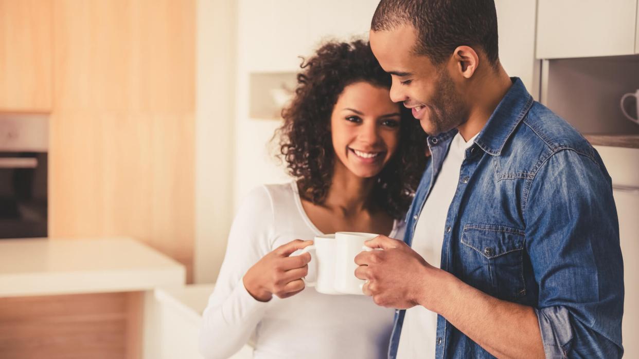 eautiful Afro American couple is drinking coffee, hugging and smiling while resting in kitchen.