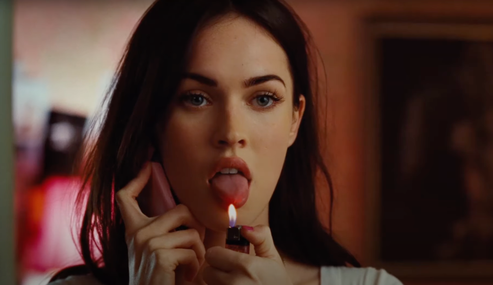 Megan Fox holds a pink phone to her ear while extending her tongue towards the flame of a lighter in a dramatic scene