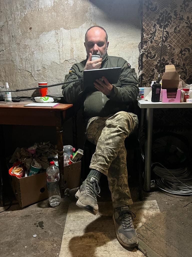 Yuriy Stetskiv speaks into a walkie-talkie in a rough-hewn command post in Bakhmut. An assortment of food and objects are strewn on tables and in boxes.