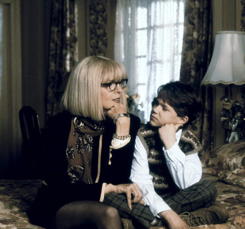 Diane Keaton in a blonde bob cut sitting on a bed next to a little boy looking at her