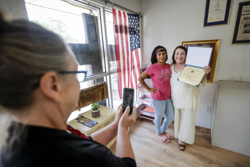 Administrative Assistant Annette Anthony, left, takes a photo of Arilya Martin, 26, second from left, and Kitty Sibley Morrison, principal and founder of Springfield Preparatory School, as Morrison holds Martin's diploma from the school in Springfield, La., Thursday, July 27, 2023. Nearly 9,000 private schools in Louisiana don’t need state approval to grant degrees. Non-approved schools make up a small percentage of the state total. But the students in Louisiana’s off-the-grid school system are a rapidly growing example of the national fallout from COVID-19 — families disengaging from traditional education. (AP Photo/Scott Threlkeld)