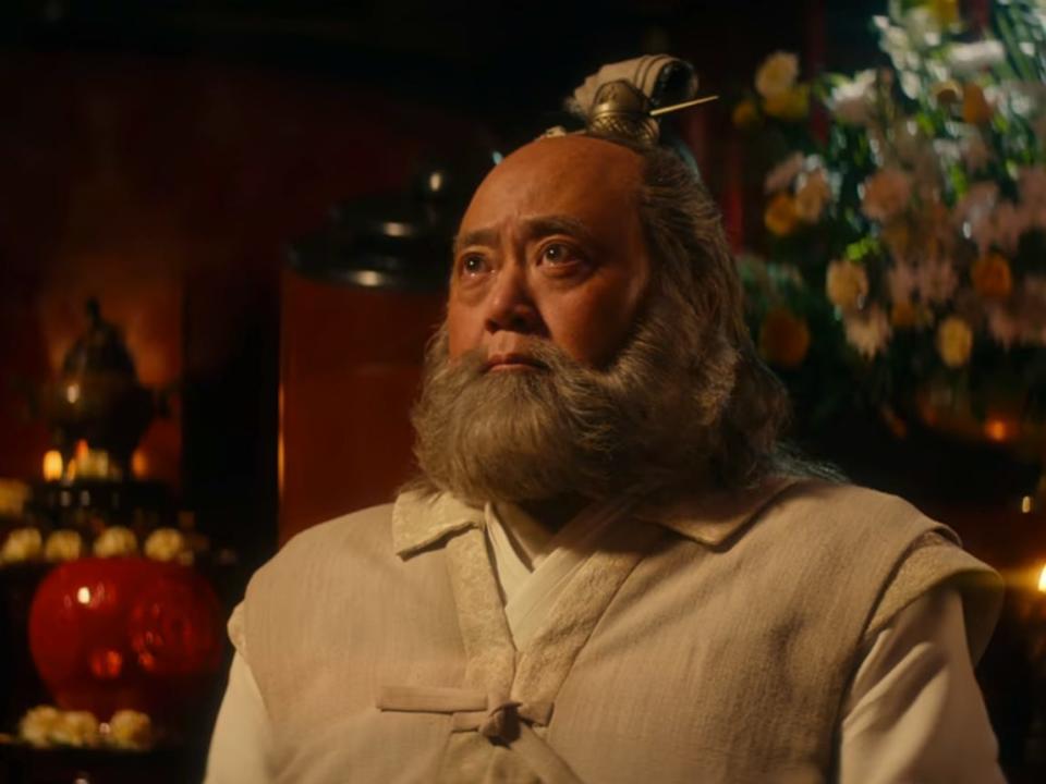 iroh in the live action avatar, wearing white and beige robes and looking up emotionally. there are flowers for a memorial service around him