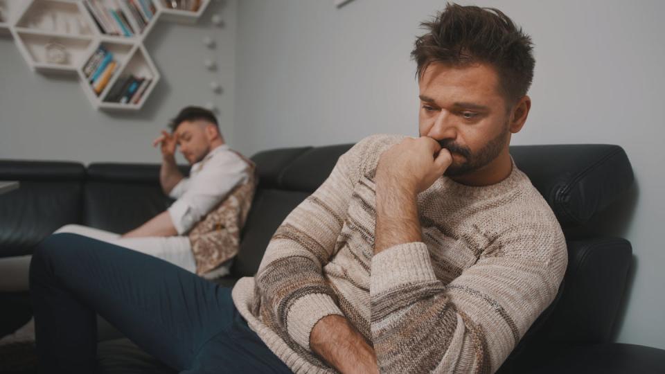 Two men sit apart on a couch with sad expressions