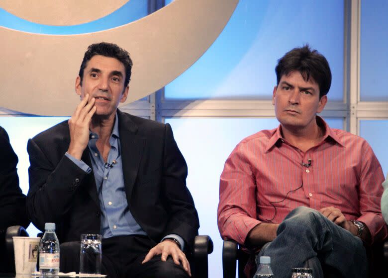 Chuck Lorre in black suit with blue shirt and Charlie Sheen in salmon shirt, seated, talking from stage