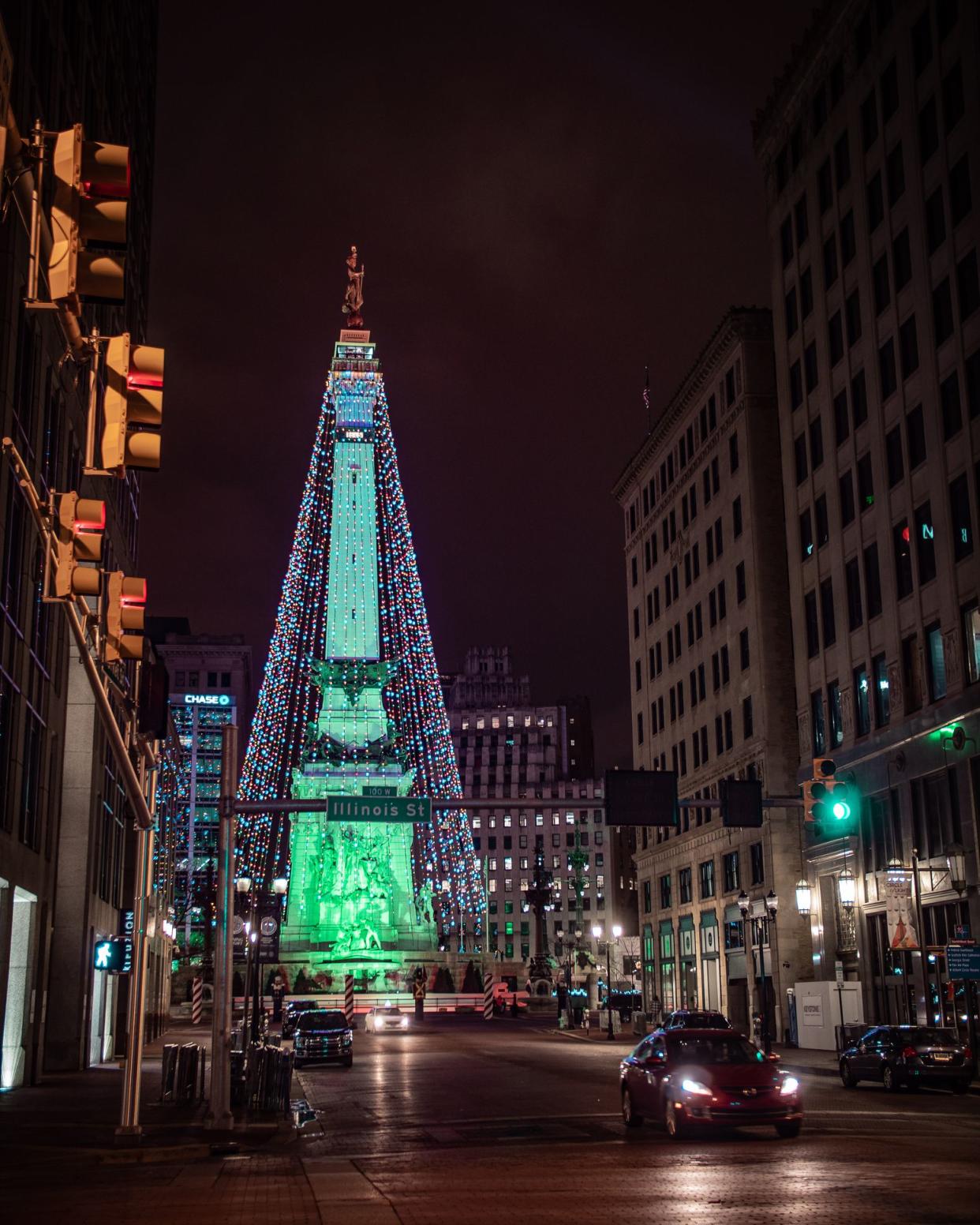 Large lit Christmas tree Fountain Square, Downtown Indianapolis, lights lit at night, with a car driving on a road