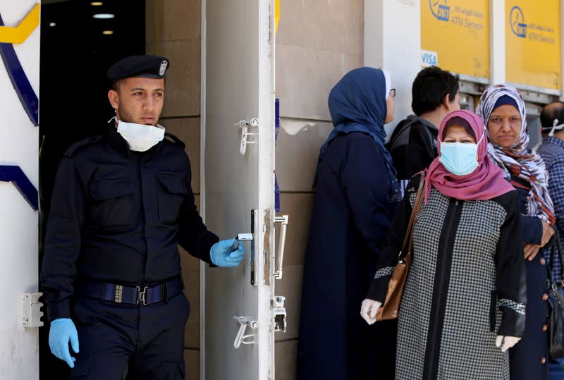 FILE PHOTO: Palestinian policeman stands guard as a woman wearing a mask waits with other people outside a bank to withdraw cash, in Gaza City