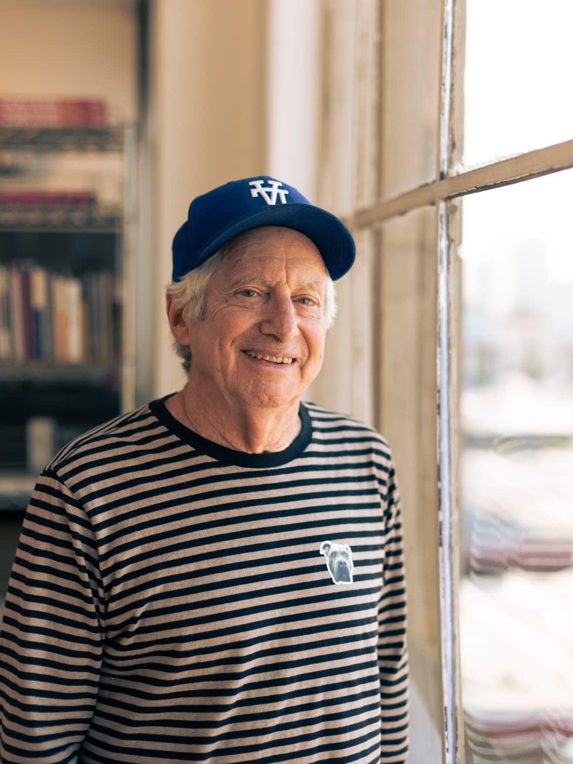 Robbie Conal stands by a window in a striped black and white shirt and a baseball cap with the letters "LA" upside down