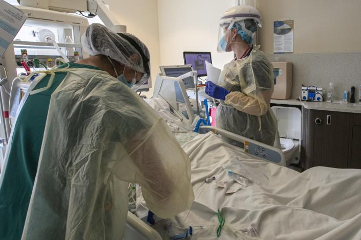 Two health workers in gowns, masks and face shields stand on either side of a patient bed