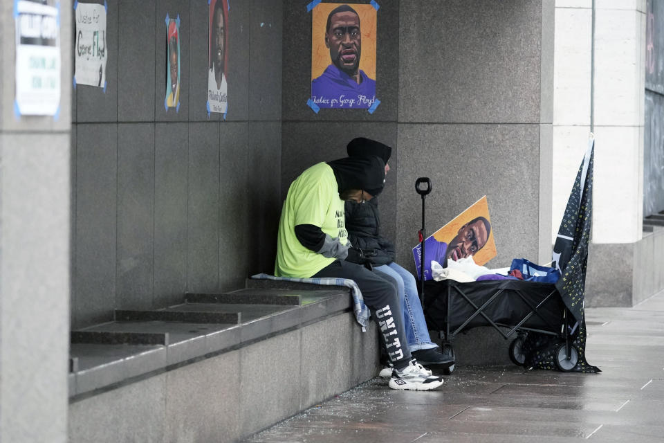 Two people with a George Floyd sign sit in a sheltered spot across the street from the Hennepin County Government Center, Wednesday, March 17, 2021, in Minneapolis where the trial for former Minneapolis police officer Derek Chauvin continues with jury selection. Chauvin is charged with murder in the death of George Floyd during an arrest last may in Minneapolis. AP Photo/Jim Mone)